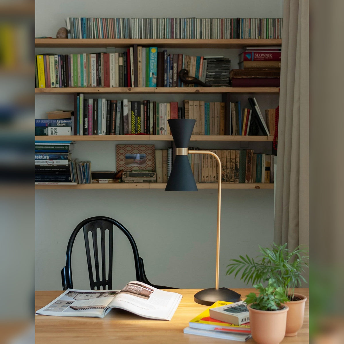 Lampe à poser Cornet noire et dorée sur un bureau en bois devant des étagères de livres