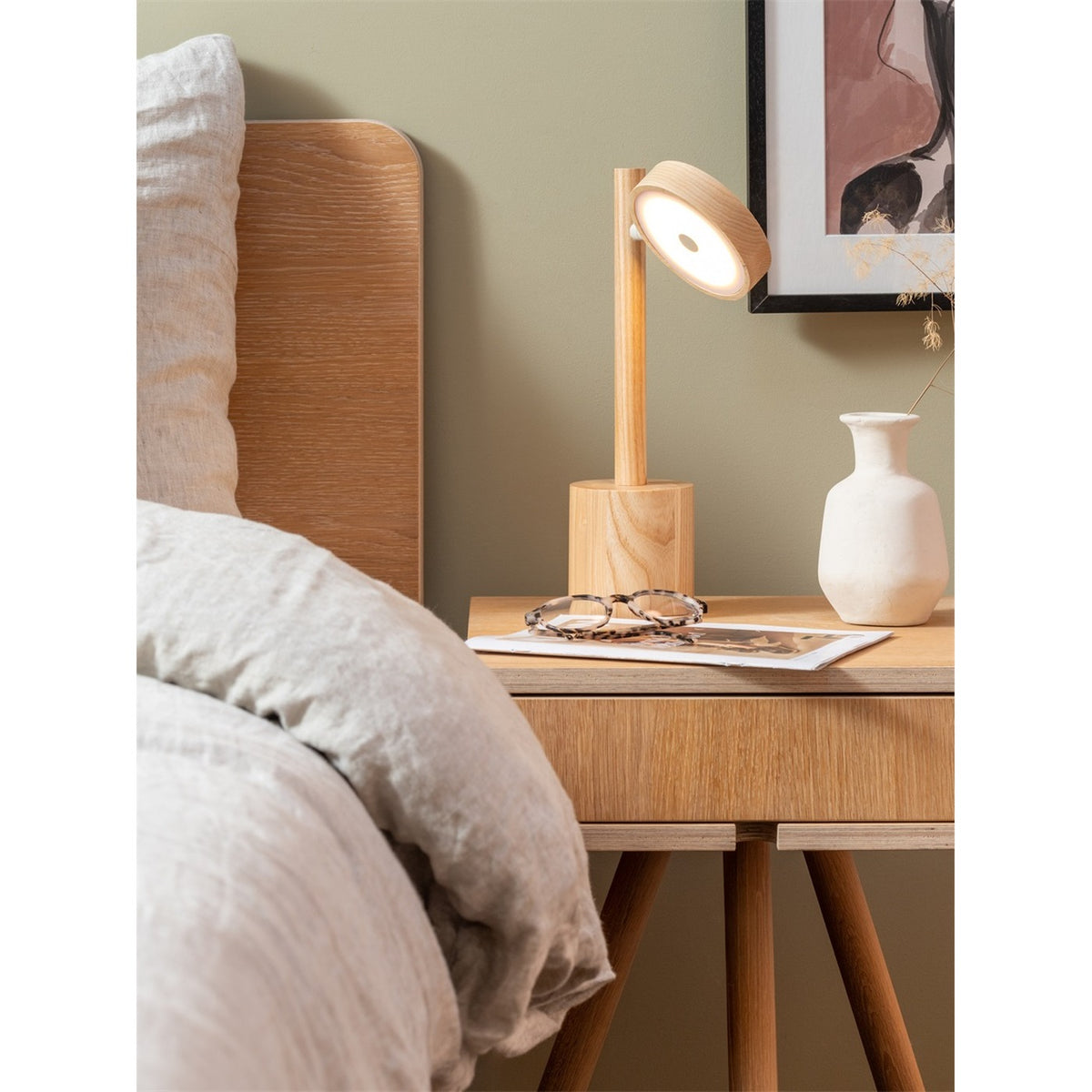 Lampe Okinawa frêne naturel allumée sur une table de chevet en bois près d'un lit en lin et d'un vase blanc