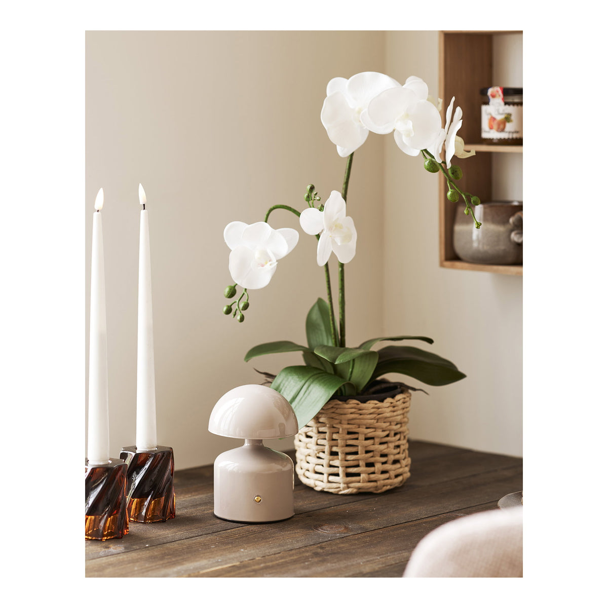 Lampe Aulla grise sur une table en bois entre deux chandelles et une orchidée blanche en cache-pot tressé