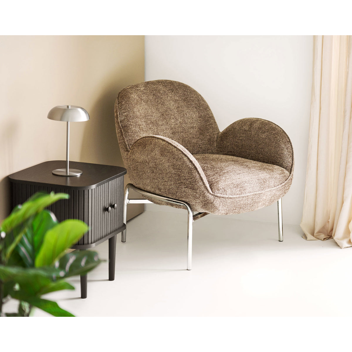 Lampe Fenwick argentée sur un chevet noir cannelé près d'un fauteuil en tissu chiné et d'une plante