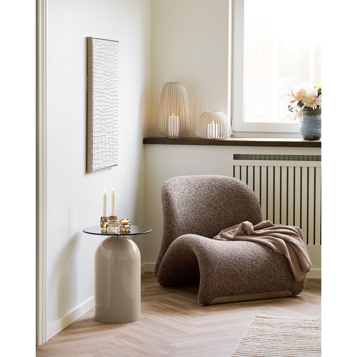 Lanterne Stoense beige allumée sur un rebord de fenêtre en bois sombre près d'une lanterne plus haute et d'un fauteuil en bouclé