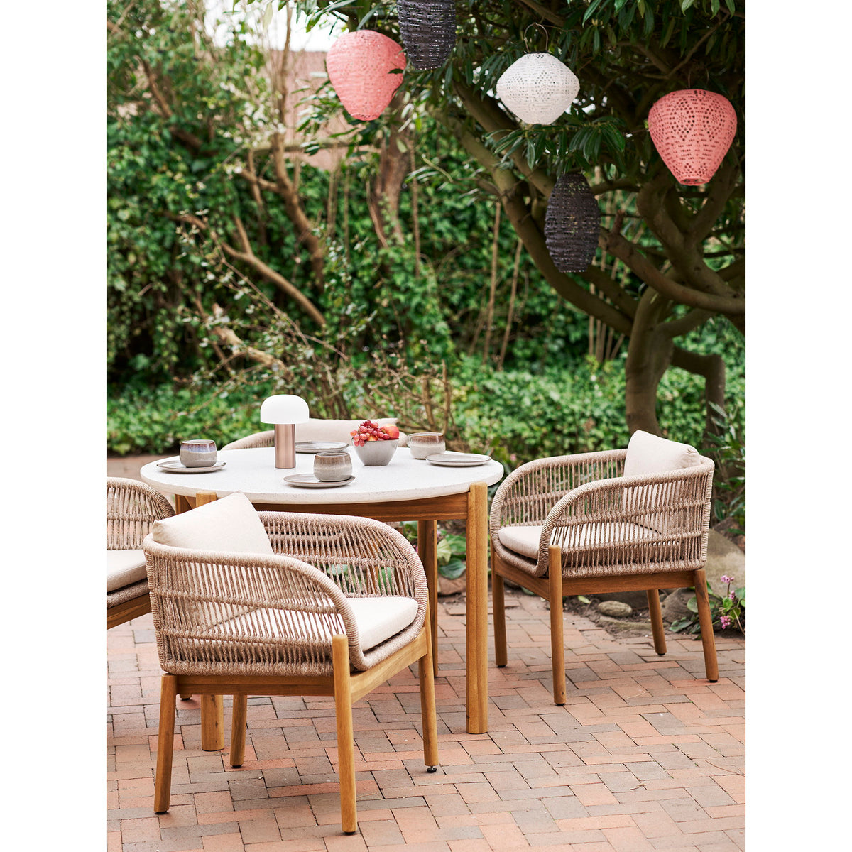 Deux lanternes Greatham rose poudré suspendues dans un arbre avec d'autres modèles de lanternes au-dessus d'une table de terrasse