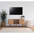Meuble TV Nikko pin naturel sous un téléviseur mural à côté d'un eucalyptus en panier tressé sur un parquet à chevrons