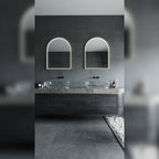 Deux miroirs lumineux en arche allumés en lumière blanche au-dessus de deux vasques en verre posées sur un plan en pierre grise