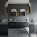 Deux miroirs lumineux en arche allumés en lumière chaude au-dessus de deux vasques en verre posées sur un plan en pierre grise