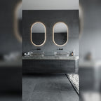 Deux miroirs lumineux ovales allumés en lumière chaude au-dessus de vasques en verre sur un plan en pierre grise et un meuble en bois sombre