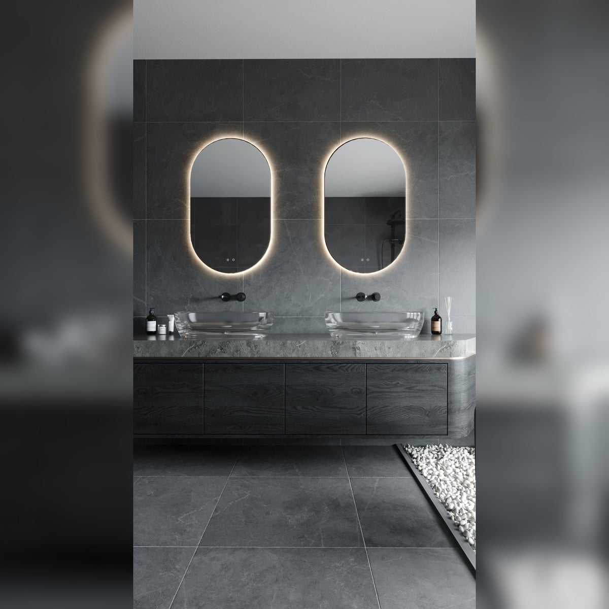 Deux miroirs lumineux ovales allumés en lumière chaude au-dessus de vasques en verre sur un plan en pierre grise et un meuble en bois sombre