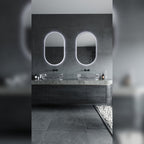 Deux miroirs lumineux ovales allumés en lumière blanche froide au-dessus de vasques en verre dans une salle de bain en pierre grise