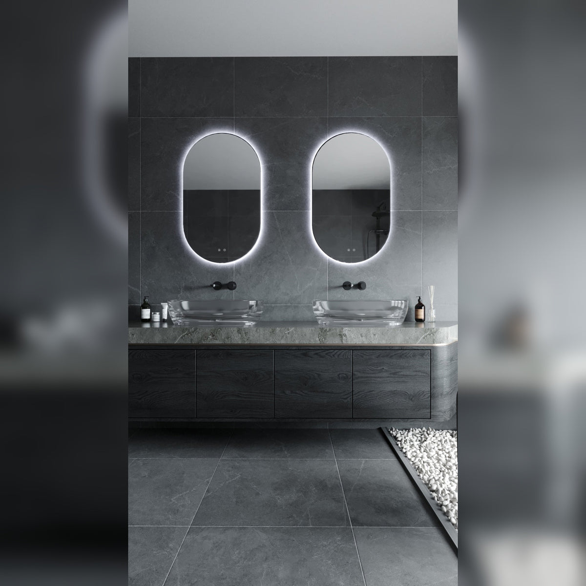 Deux miroirs lumineux ovales allumés en lumière blanche froide au-dessus de vasques en verre dans une salle de bain en pierre grise