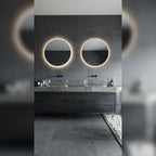 Deux miroirs lumineux ronds au halo blanc chaud au-dessus d'une double vasque en verre dans une salle de bains en pierre grise