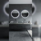 Deux miroirs lumineux ronds au halo blanc froid au-dessus d'une double vasque en verre dans une salle de bains en pierre grise