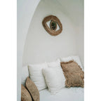 Miroir Raffia Eye dans une alcôve blanche au-dessus d'une banquette garnie de coussins en raphia et en coton