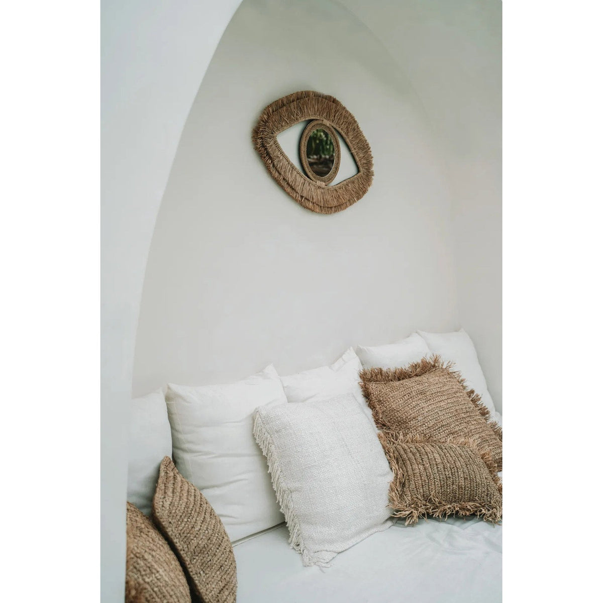 Miroir Raffia Eye dans une alcôve blanche au-dessus d'une banquette garnie de coussins en raphia et en coton