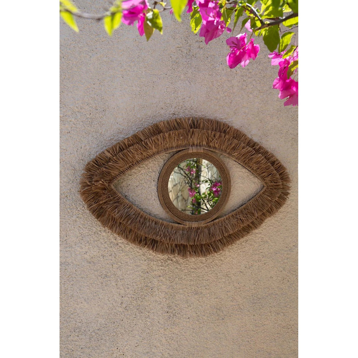 Miroir Raffia Eye en raphia accroché sur un mur en enduit beige sous des fleurs de bougainvillier