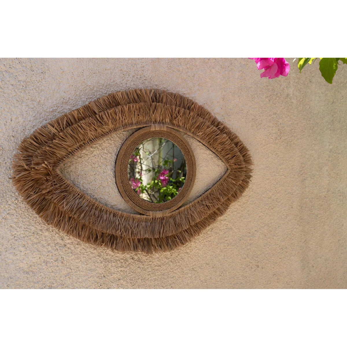 Miroir Raffia Eye en raphia sur un mur en enduit beige reflétant un bougainvillier en fleurs