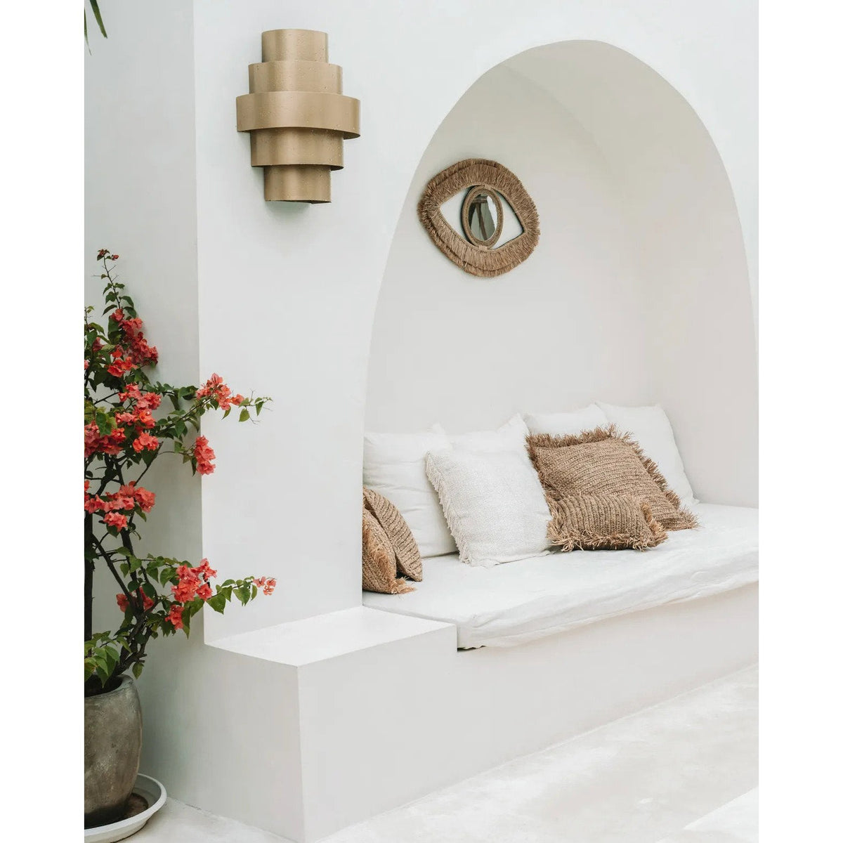 Miroir Raffia Eye dans une niche blanche en arc au-dessus d'une banquette près d'un bougainvillier en pot