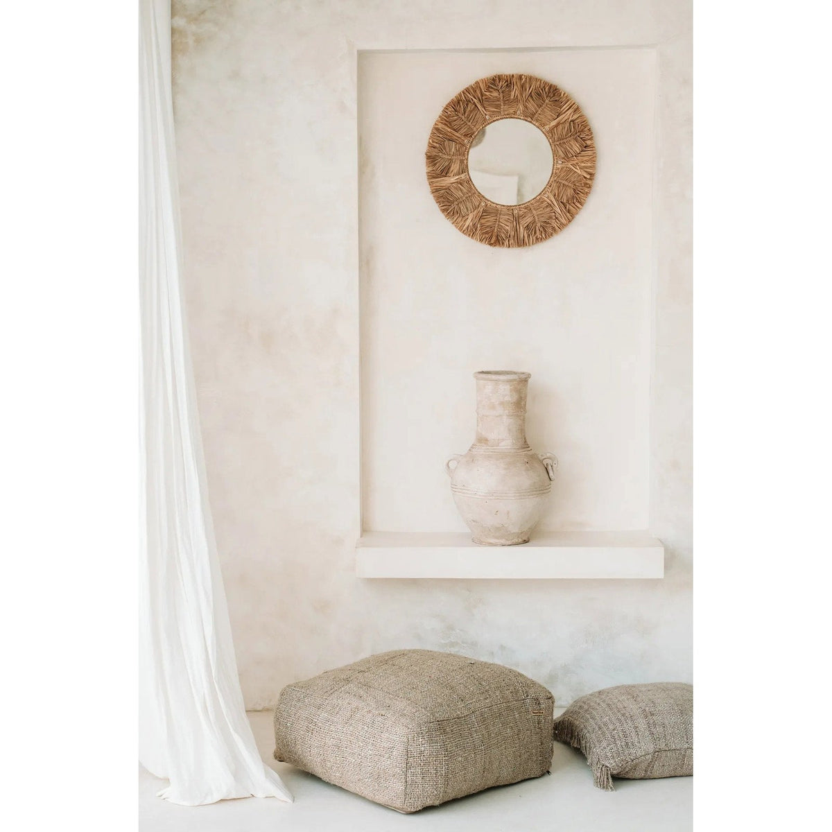 Miroir Palm Tree en raphia dans une niche blanche au-dessus d'une jarre en terre avec un pouf et un coussin au sol