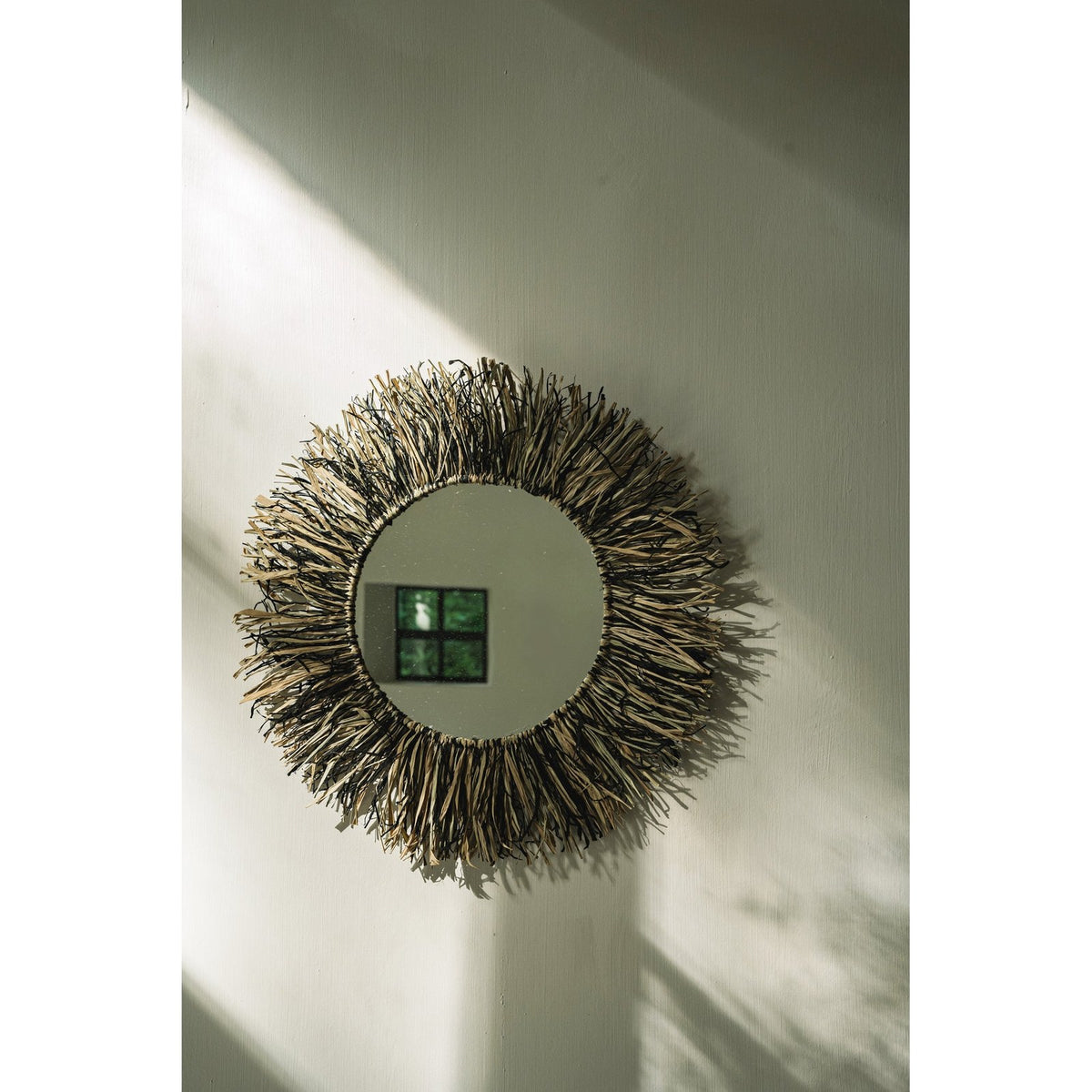 Miroir Raffia Fringe accroché sur un mur clair dans un rai de lumière avec son ombre portée