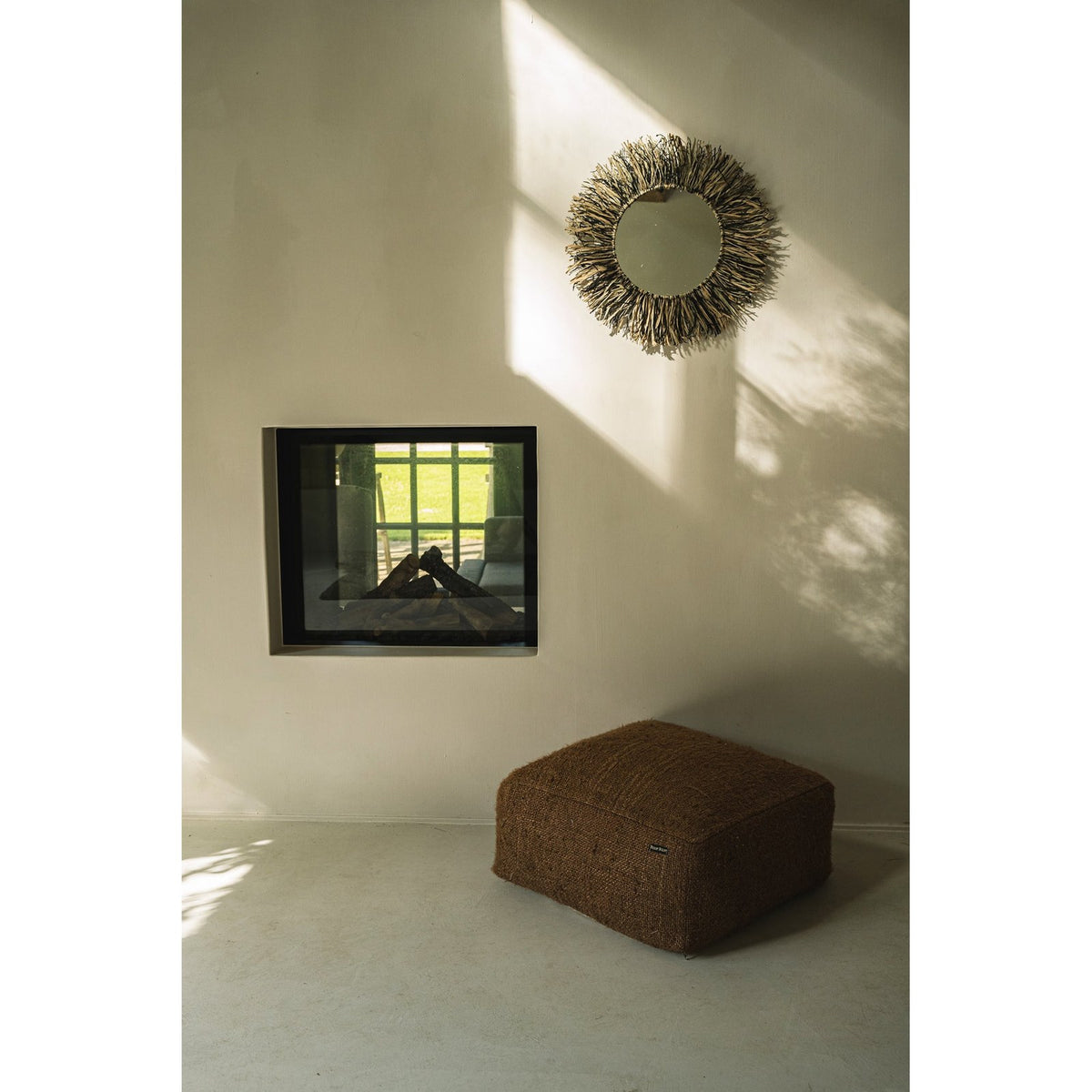 Miroir Raffia Fringe en raphia naturel et noir sur un mur blanc au-dessus d'un pouf près d'une cheminée à foyer vitré