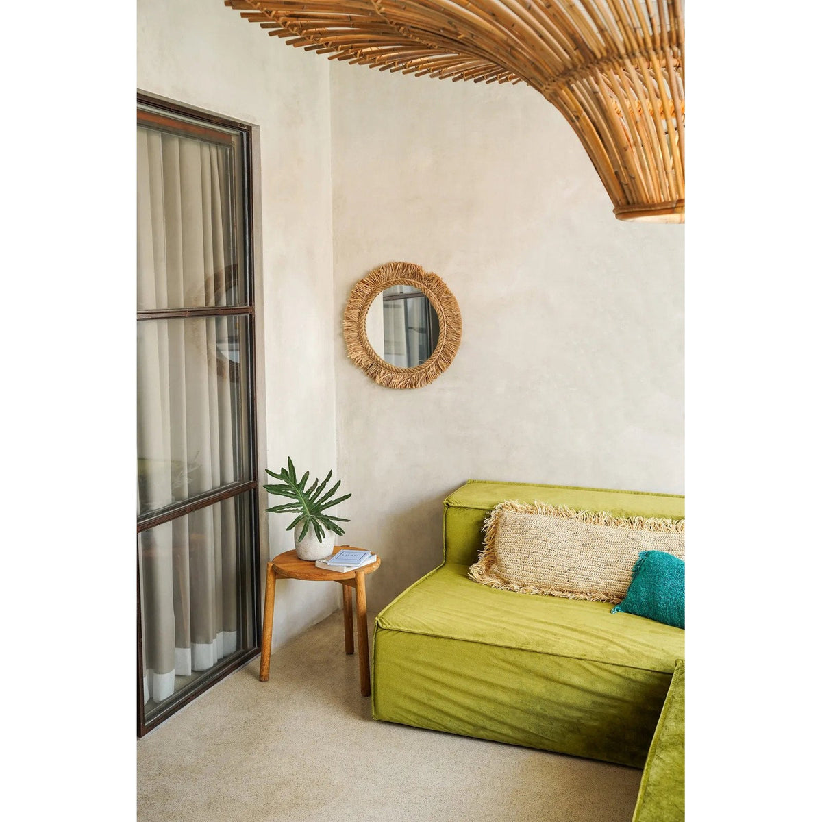 Miroir Tahiti sur un mur clair de salon entre une porte vitrée et un canapé vert sous une suspension en rotin