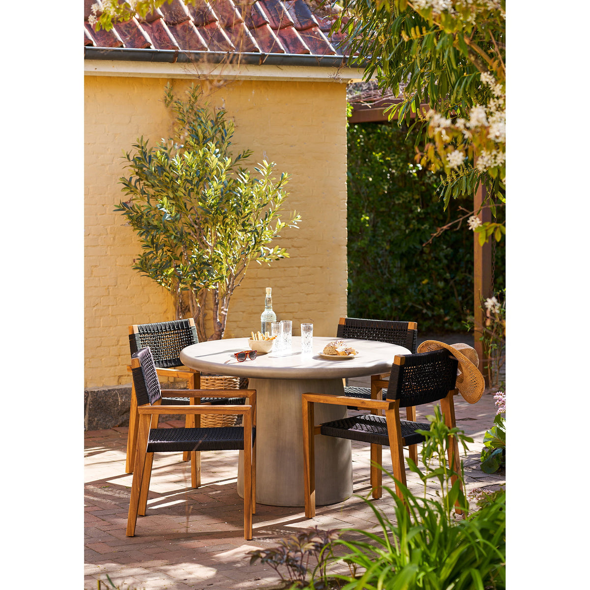Terrasse avec table ronde et fauteuils en bois et corde noire devant un olivier artificiel dans un panier