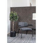 Olivier artificiel dans un panier tressé noir près d'un fauteuil gris devant un mur en briques