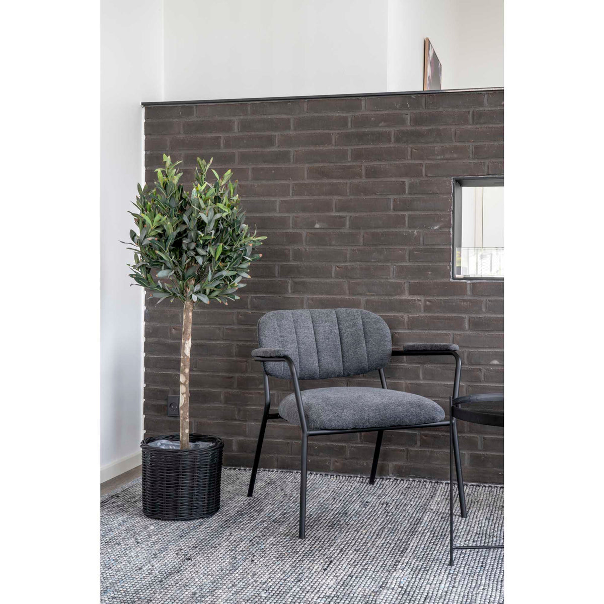 Olivier artificiel dans un panier tressé noir près d'un fauteuil gris devant un mur en briques
