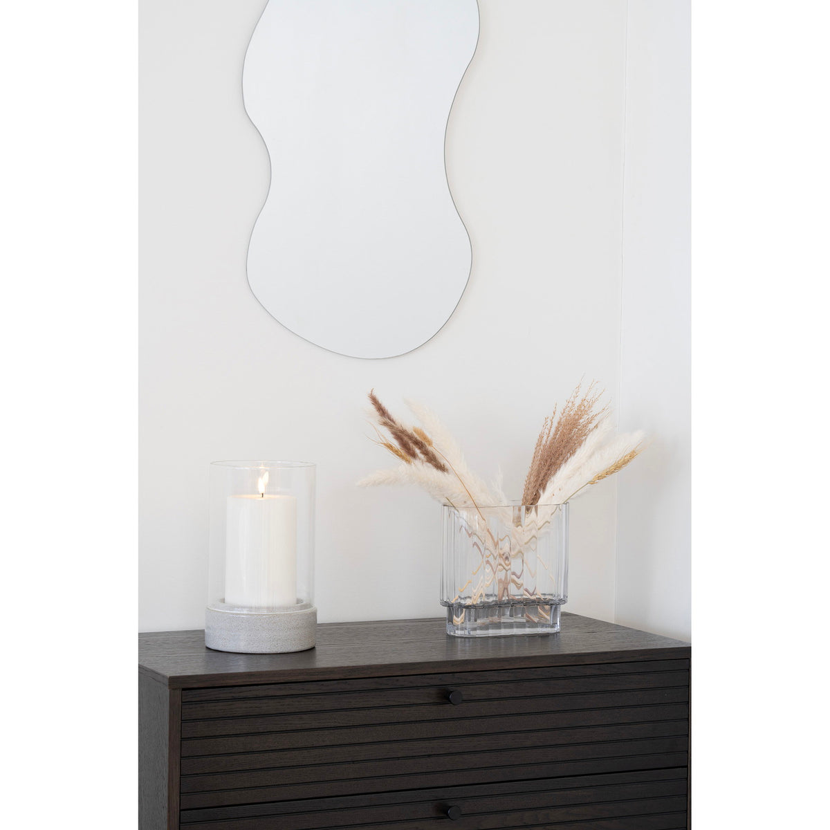 Photophore Hurricane avec une bougie pilier allumée sur une commode en bois sombre à côté d'un vase garni de pampa