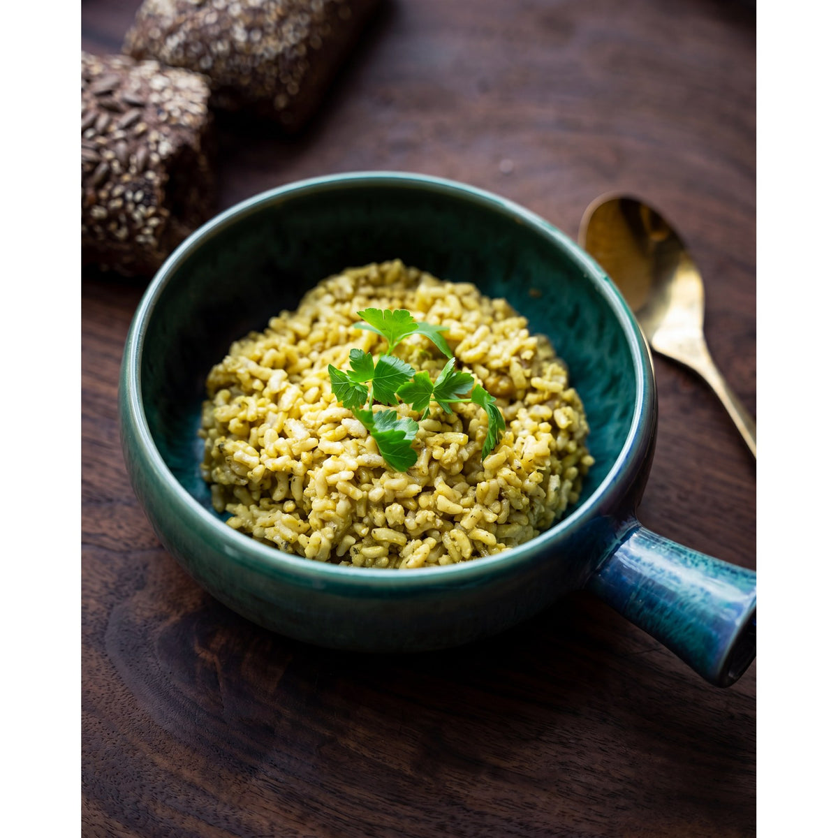 Plat à manche en grès bleu vert garni de risotto au persil sur une table en bois sombre avec une cuillère dorée