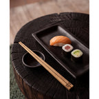 Plat à sushis 25 x 12 cm garni d'un nigiri au saumon et de deux makis sur un billot de bois sombre à côté d'une coupelle et de baguettes