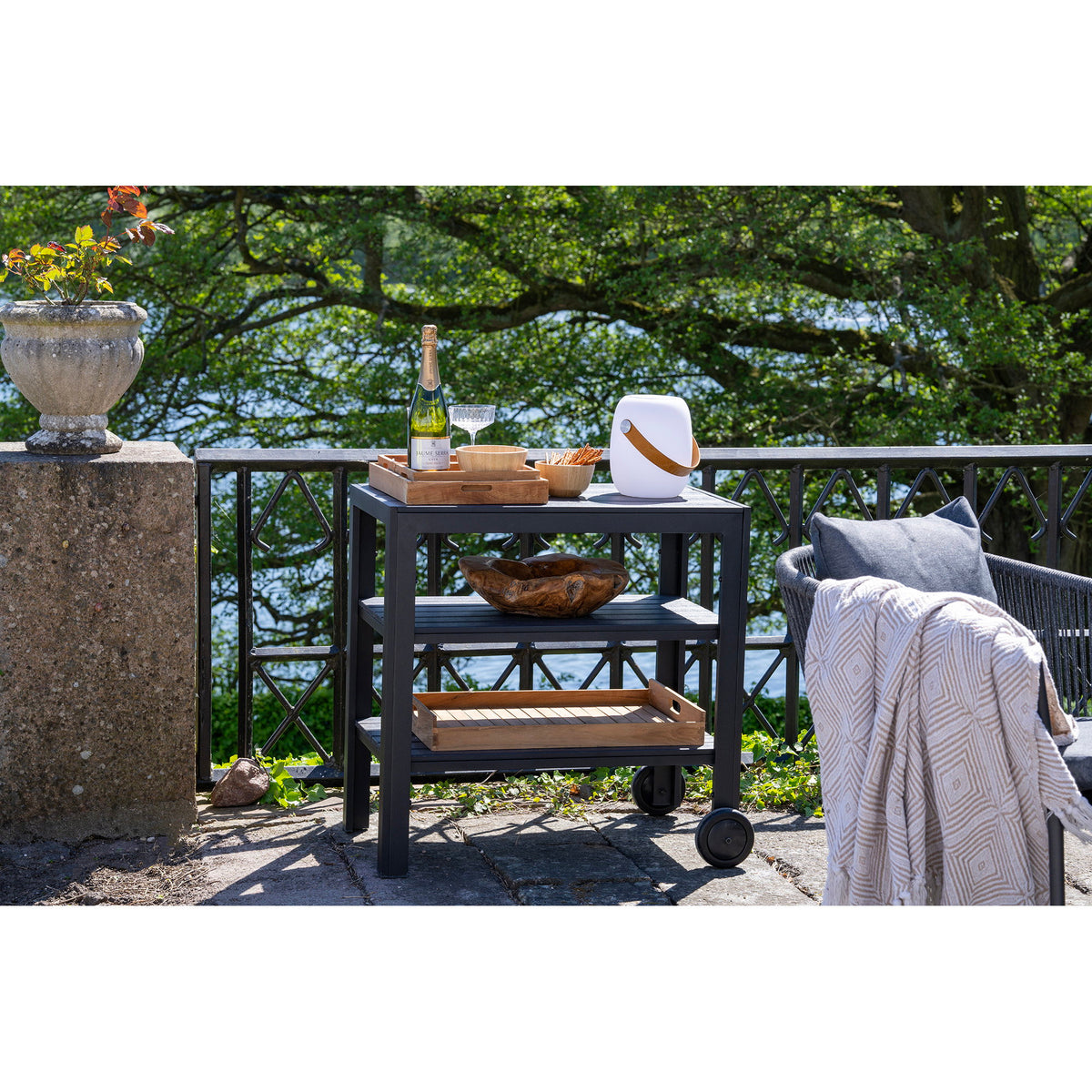 Plateaux Faro en teck sur une desserte noire de terrasse avec champagne et coupe en bois