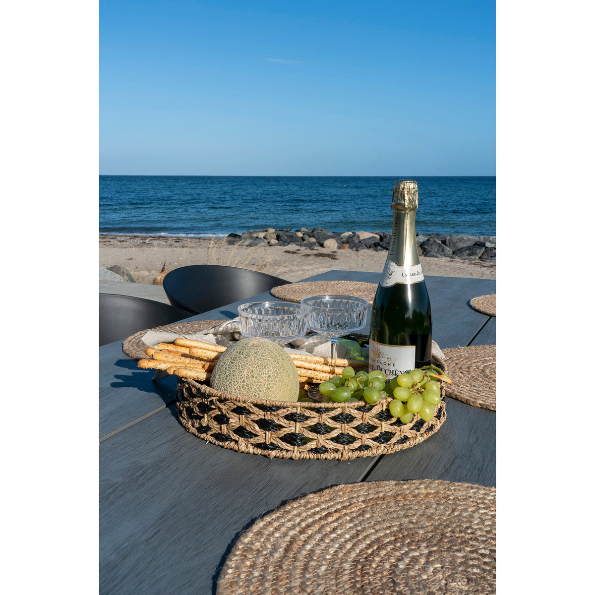 Plateau Nerja garni de melon de raisin et de gressins près d'une bouteille de champagne sur une table en bord de mer