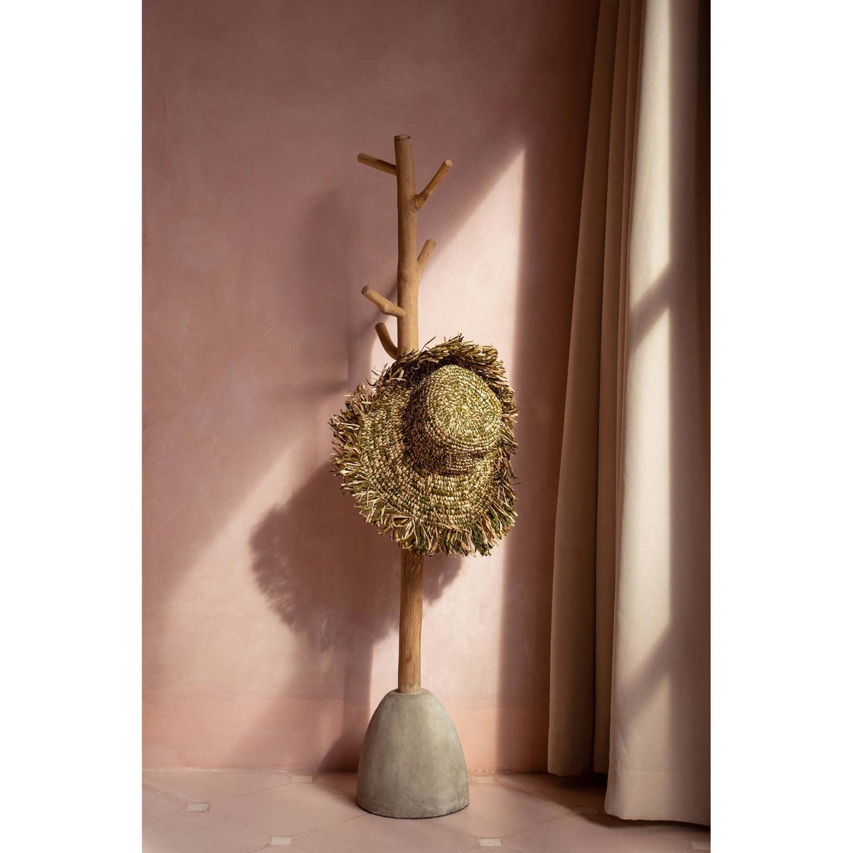 Portemanteau en teck et béton portant un chapeau de paille à franges contre un mur rose