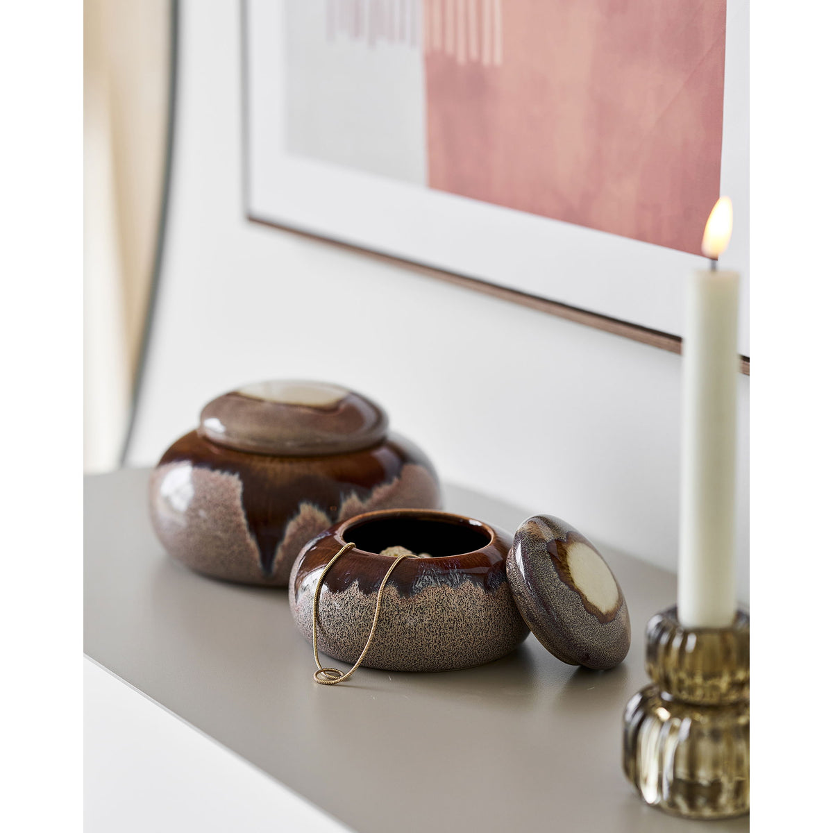 Petit pot brun chocolat ouvert avec un collier doré à côté du grand pot fermé sur une console