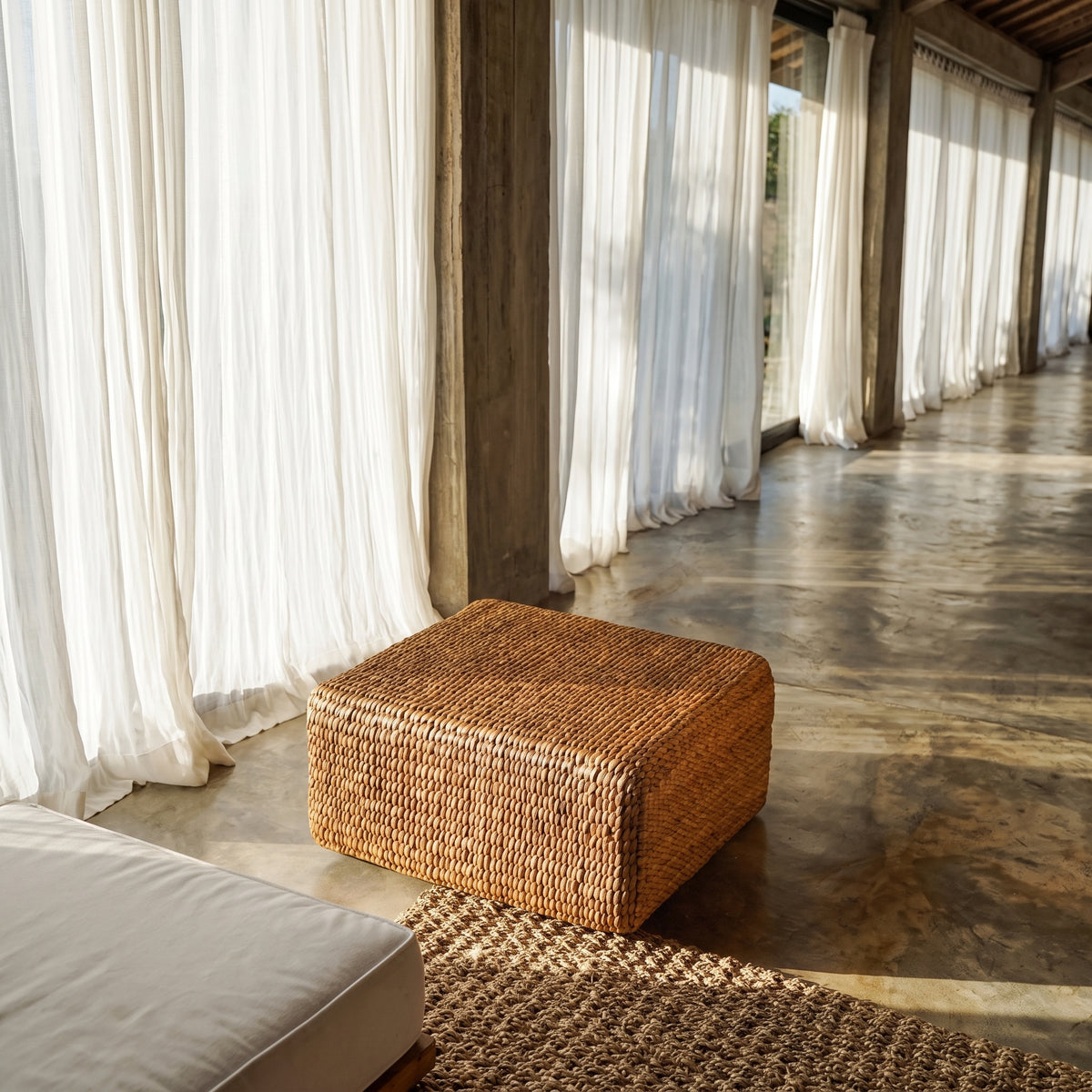 Pouf carré en jacinthe d'eau tressée près d'un tapis tressé dans une longue galerie aux voilages blancs
