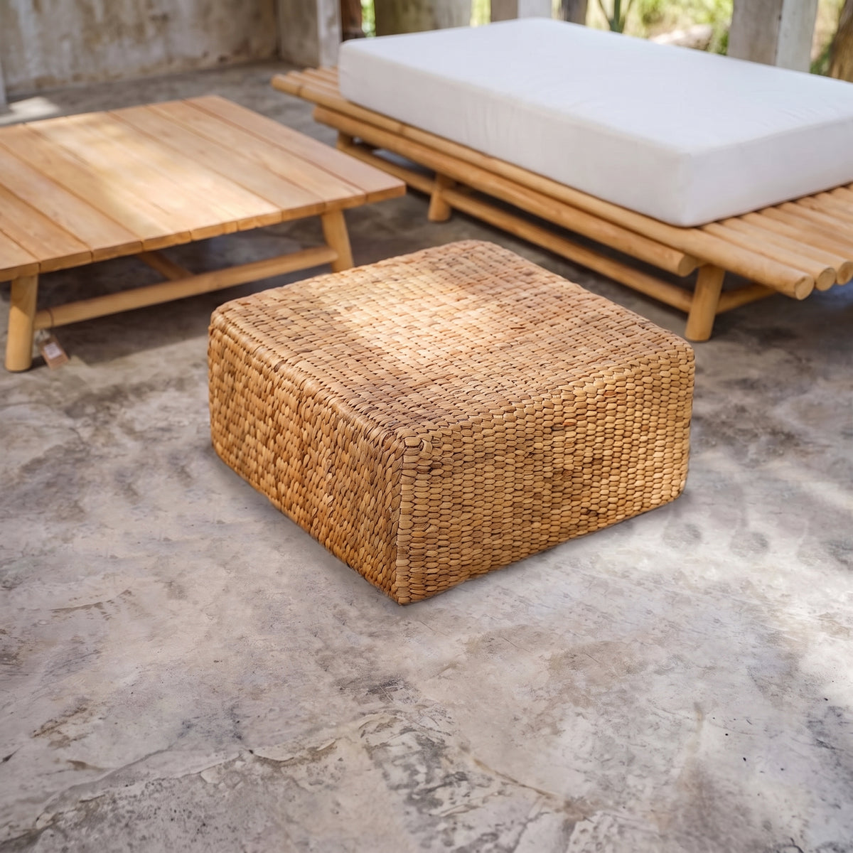 Pouf carré en jacinthe d'eau tressée entre une table basse et une banquette en bambou sur un sol en béton