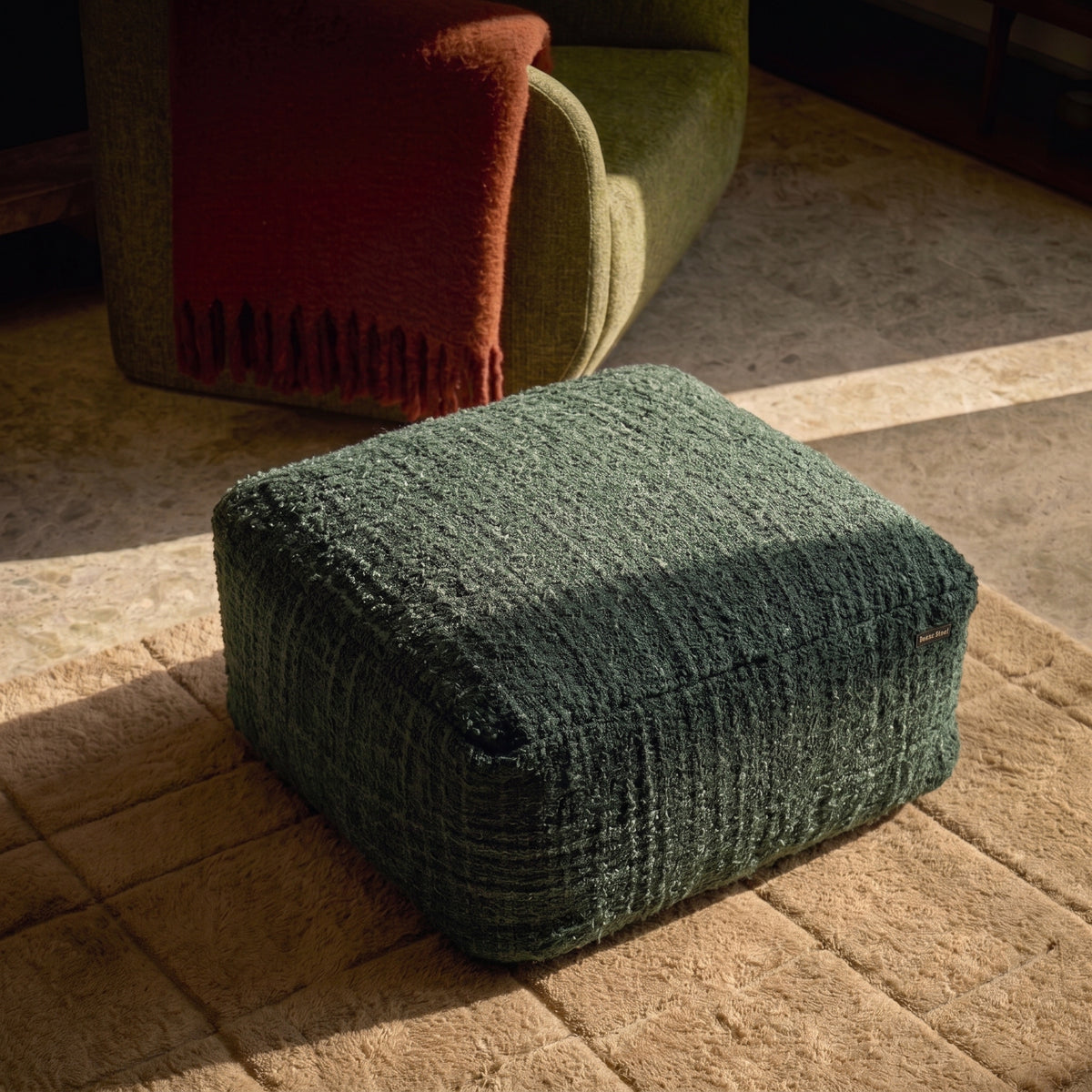 Pouf carré Oh My Gee en coton tissé vert forêt sur un tapis beige à carreaux, devant un fauteuil vert et un plaid rouille