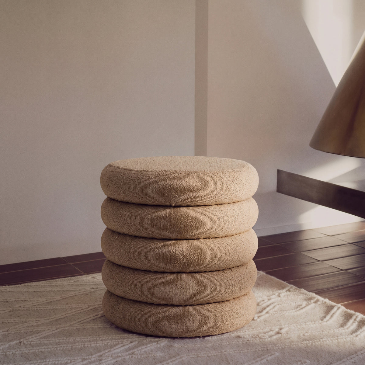 Pouf bouclé beige à bourrelets posé sur un tapis crème, à côté d'une console et d'une lampe à abat-jour laiton