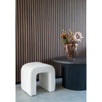 Pouf Colma blanc devant un mur en lames de bois foncé à côté d'une table basse noire et d'un vase de fleurs