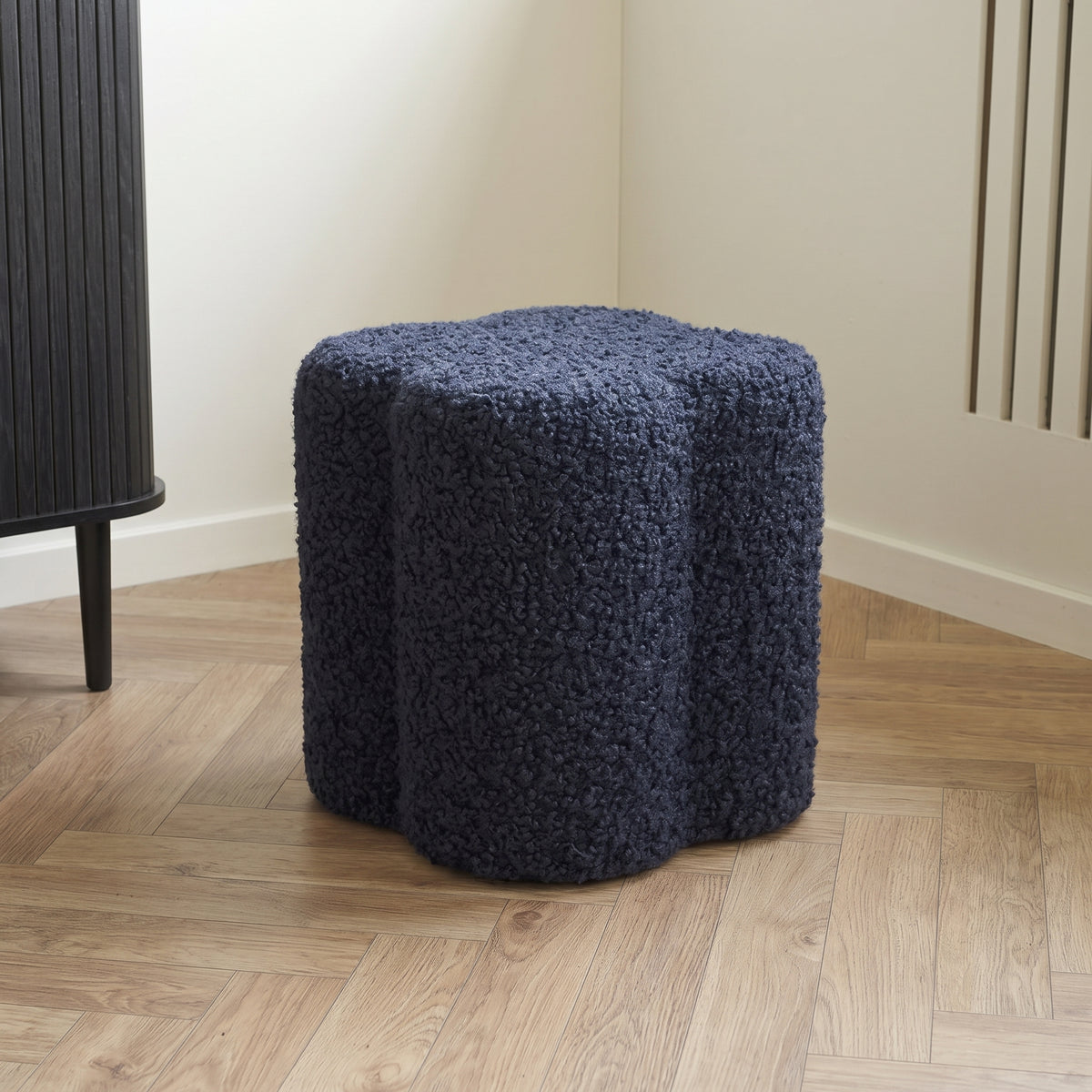 Pouf Durban bouclé bleu marine dans l'angle d'une pièce claire, sur un parquet à bâtons rompus près d'un buffet noir