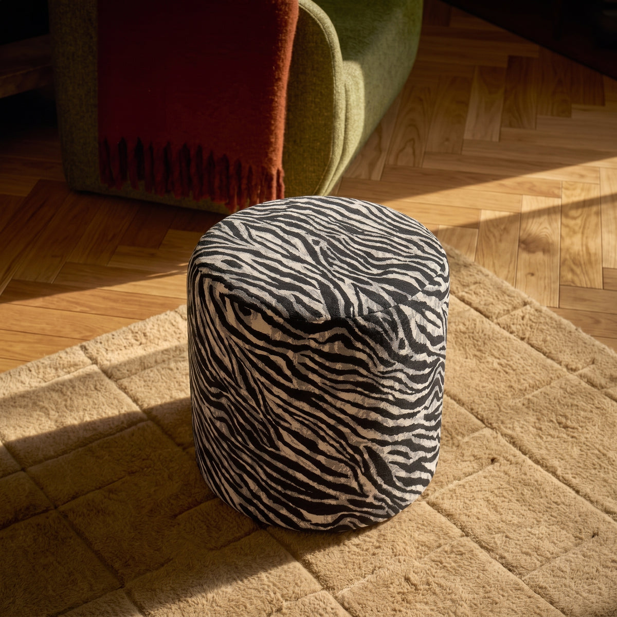 Pouf zébré noir et blanc posé sur un tapis beige devant un canapé vert, parquet en chevrons au soleil