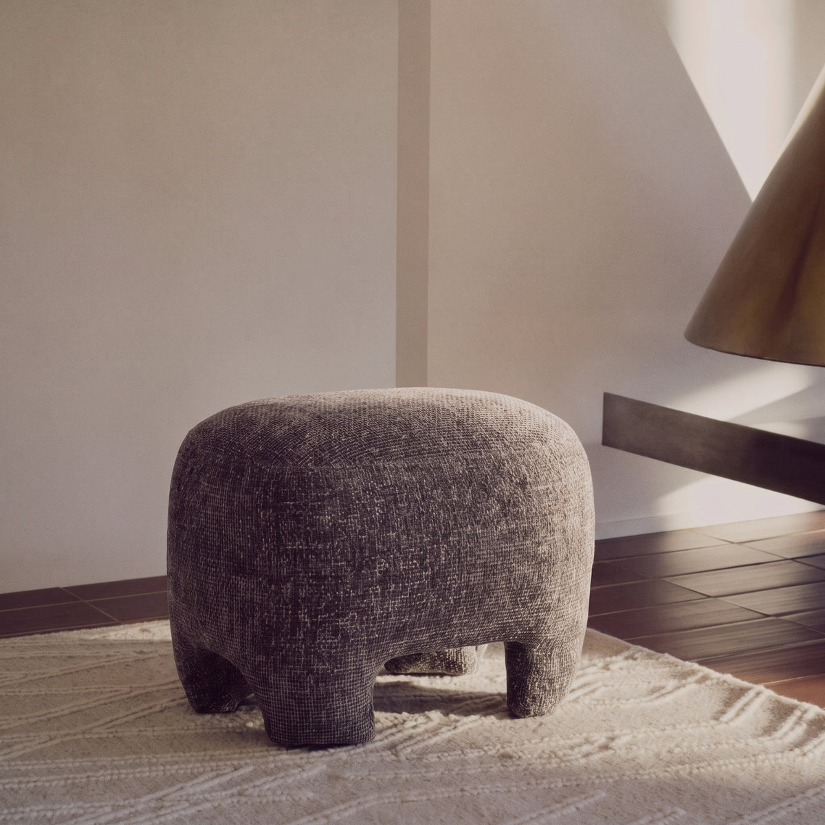 Pouf Nazare en tissu gris chiné posé sur un tapis clair, près d'une cheminée suspendue en laiton