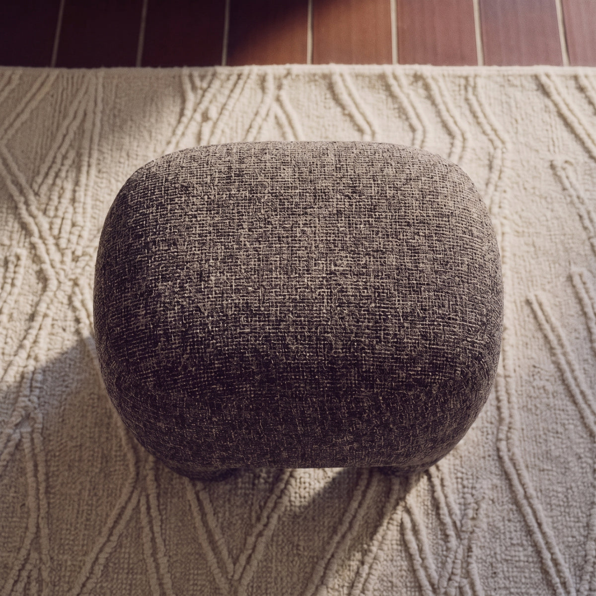 Pouf Nazare vu de dessus sur un tapis à motifs en relief, son tissu gris chiné en pleine lumière