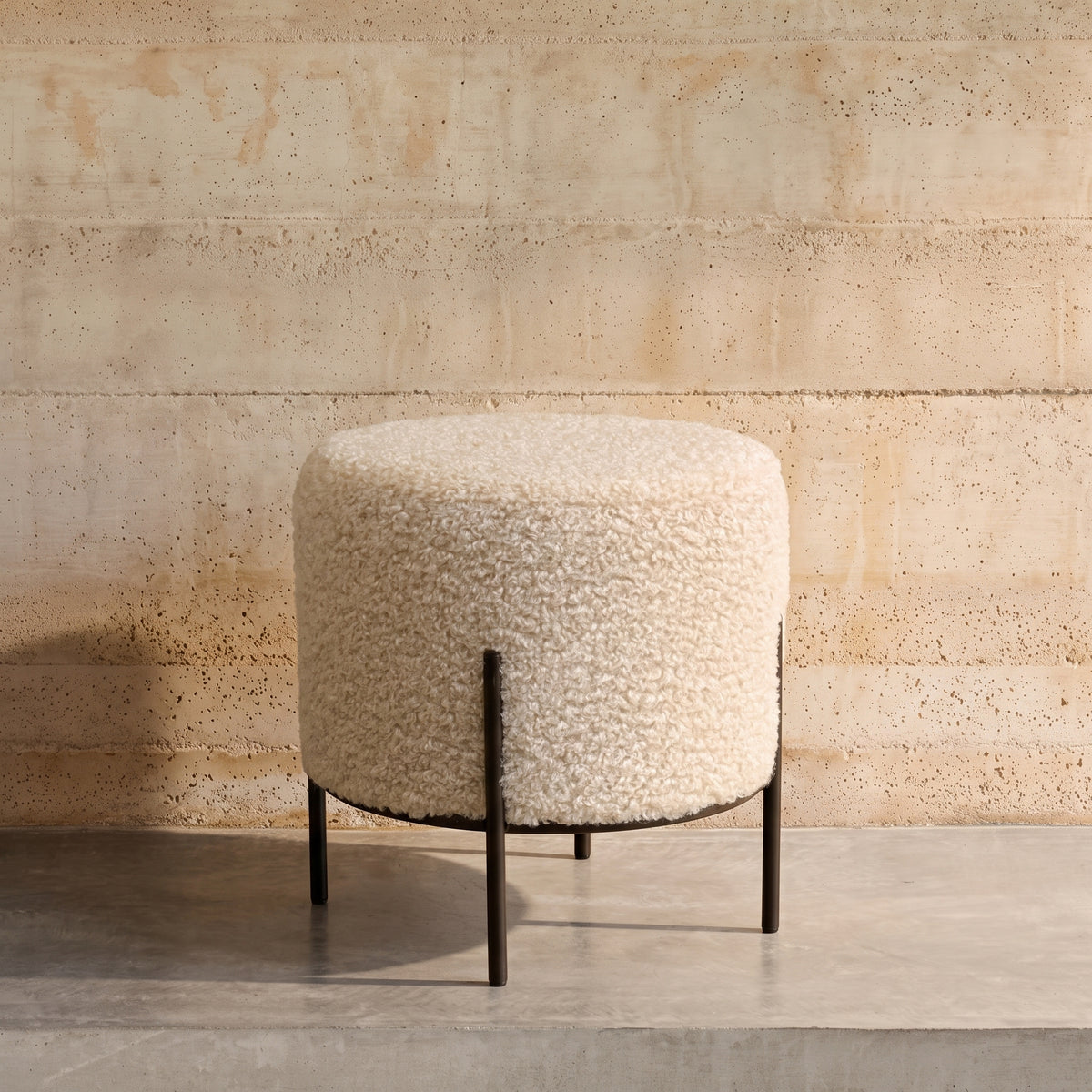Pouf rond Alford en bouclette crème devant un mur en béton banché, sur un sol en béton