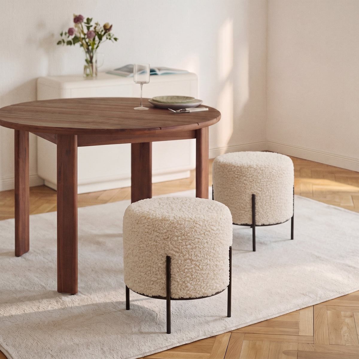 Deux poufs ronds Alford en bouclette crème près d'une table ronde en bois, sur un tapis clair et un parquet à bâtons rompus