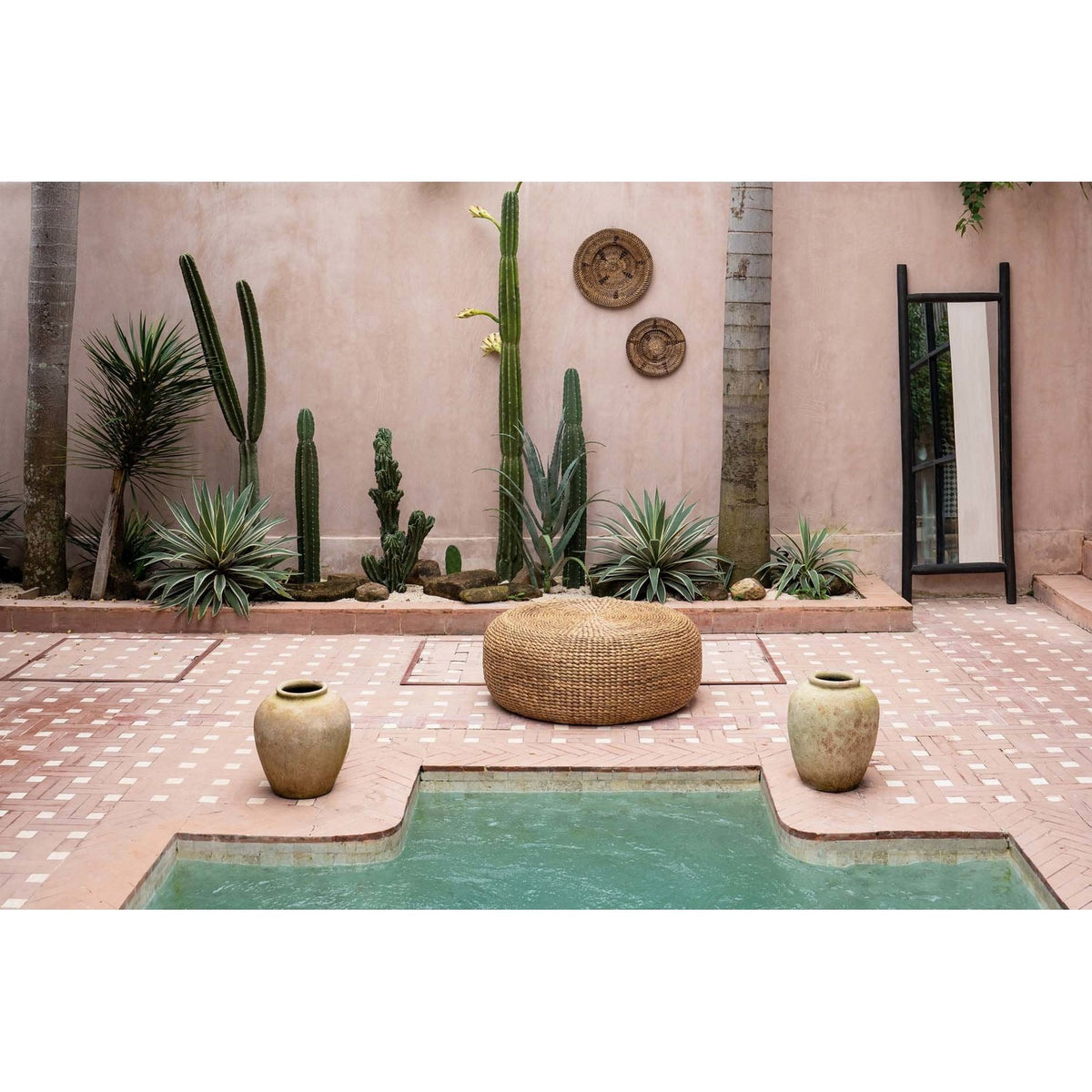 Pouf rond en jacinthe d'eau tressée au centre d'un patio carrelé entre deux jarres et des cactus
