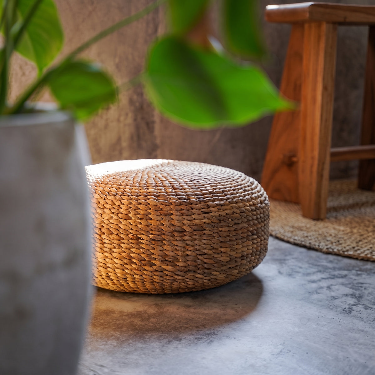 Pouf rond en jacinthe d'eau tressée sur un sol en béton près d'un tabouret en bois posé sur un tapis tressé
