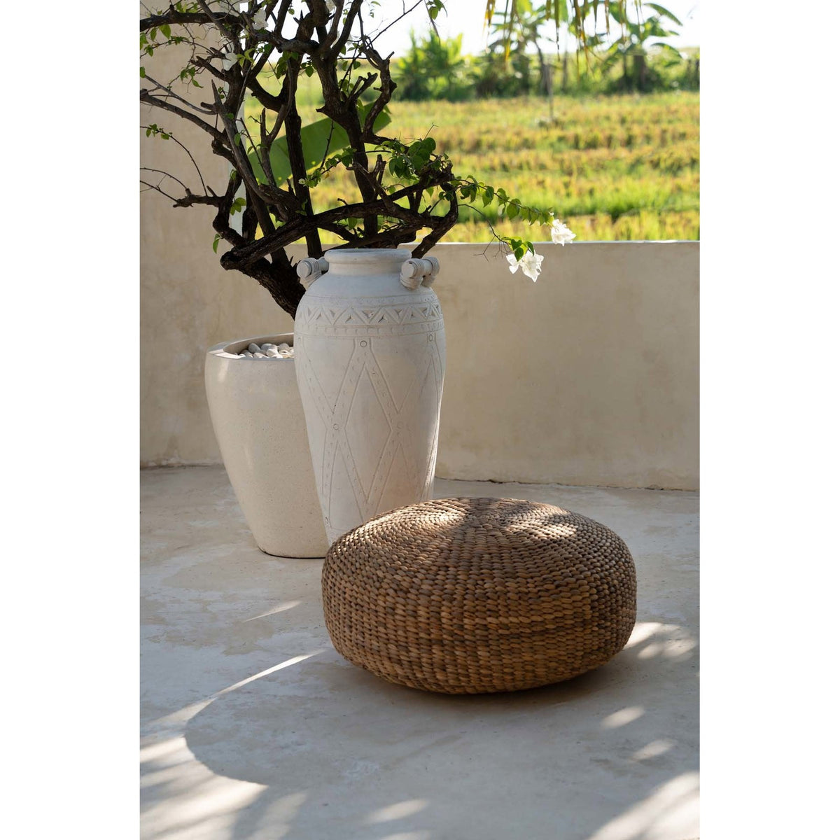 Pouf rond en jacinthe d'eau tressée sur une terrasse devant une jarre blanche et un arbuste en pot