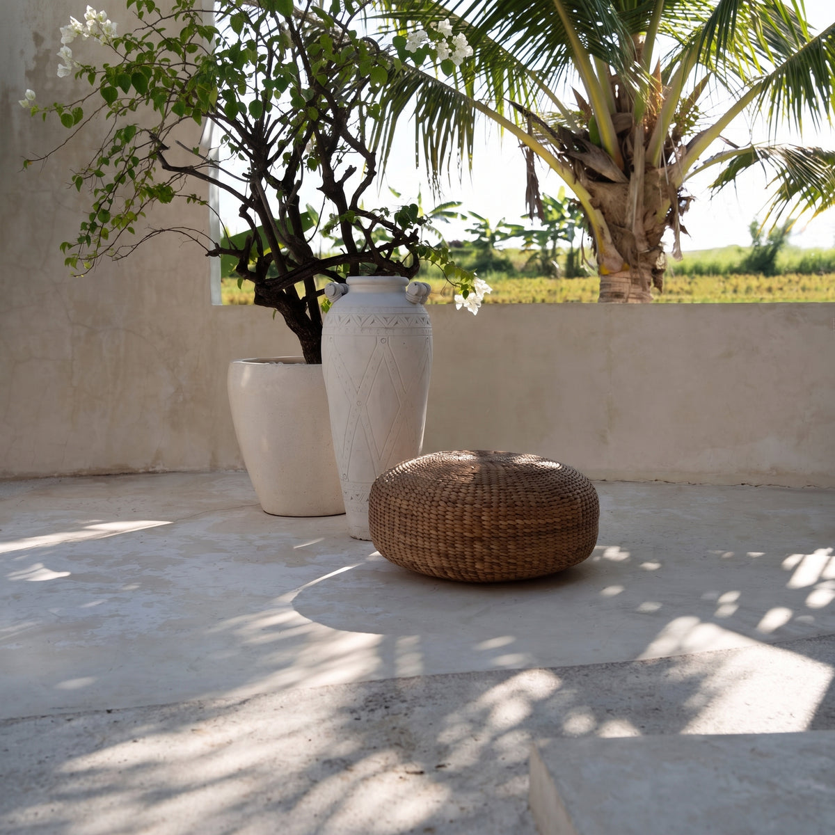Pouf rond en jacinthe d'eau tressée sur une terrasse claire près d'une jarre blanche et d'un arbuste en pot