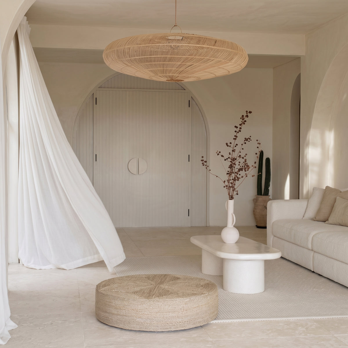 Pouf bas en jonc de mer tressé dans un salon blanc à arches, sous une grande suspension en rotin